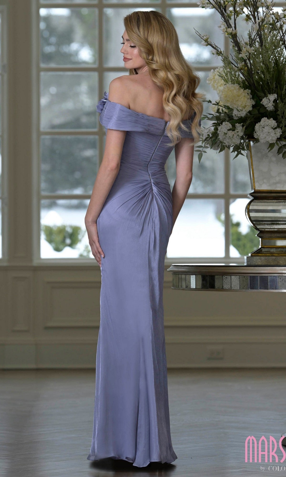 A woman with long wavy blonde hair stands indoors facing away, wearing the Marsoni M353 fitted off-the-shoulder lavender formal dress with 3D floral accents; large windows enhance the elegant atmosphere.